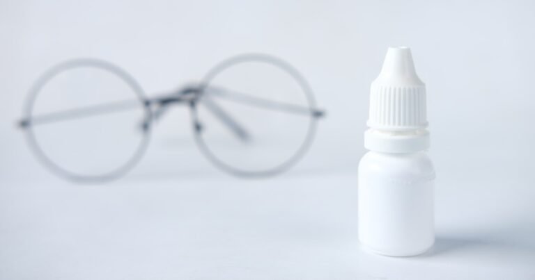 Flacon de collyre en plastique blanc, net au premier plan ; derrière, floue, une paire de lunettes rondes à fine monture métallique, sur fond blanc
