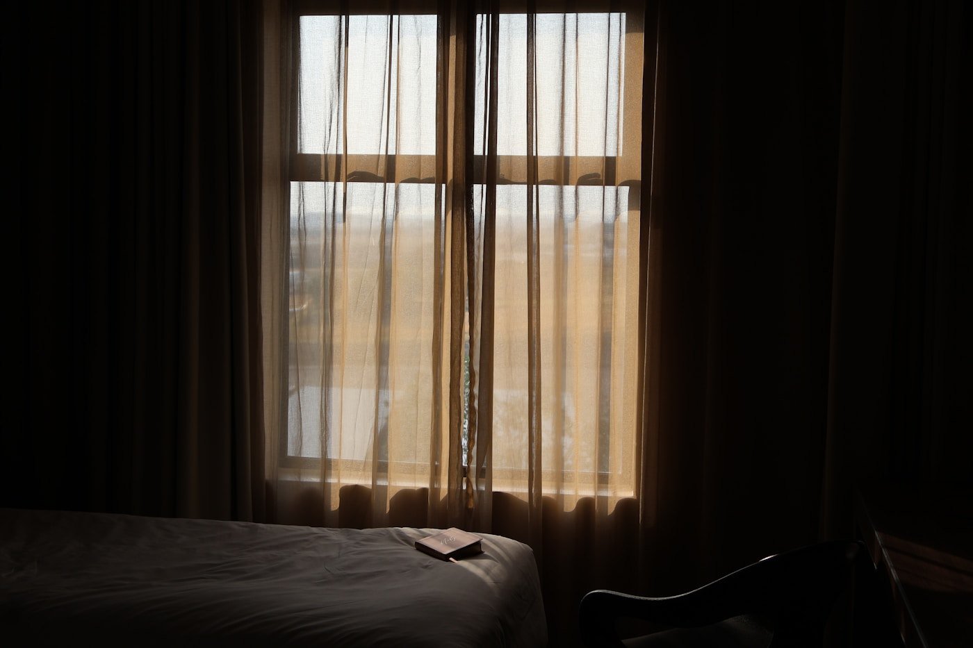 Room in half-light crossed by light filtered through a curtain, a book lying on the bed