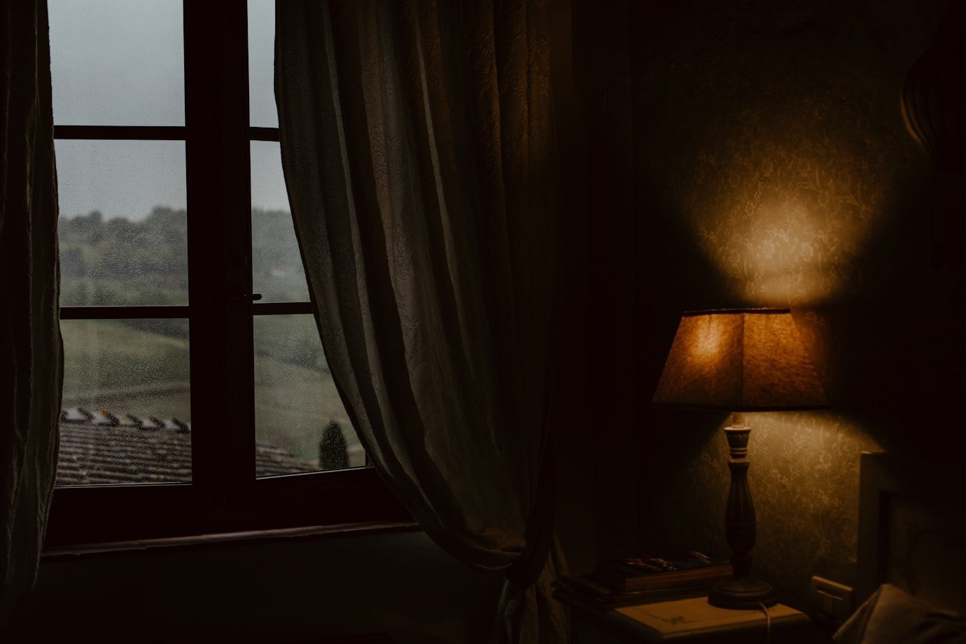 Dark room at the end of the day, grey light at the window and a bedside lamp switched on