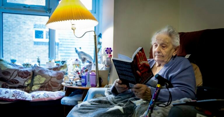 Ältere Frau liest im Sessel neben einer Lampe ein Buch